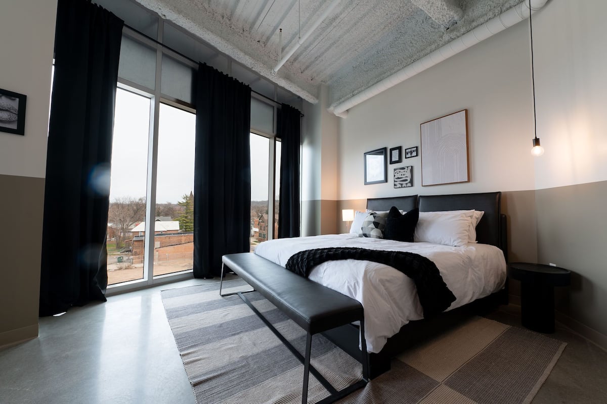 A modern hotel room with a large bed, a bench, a bedside table, and a view of the outside.
