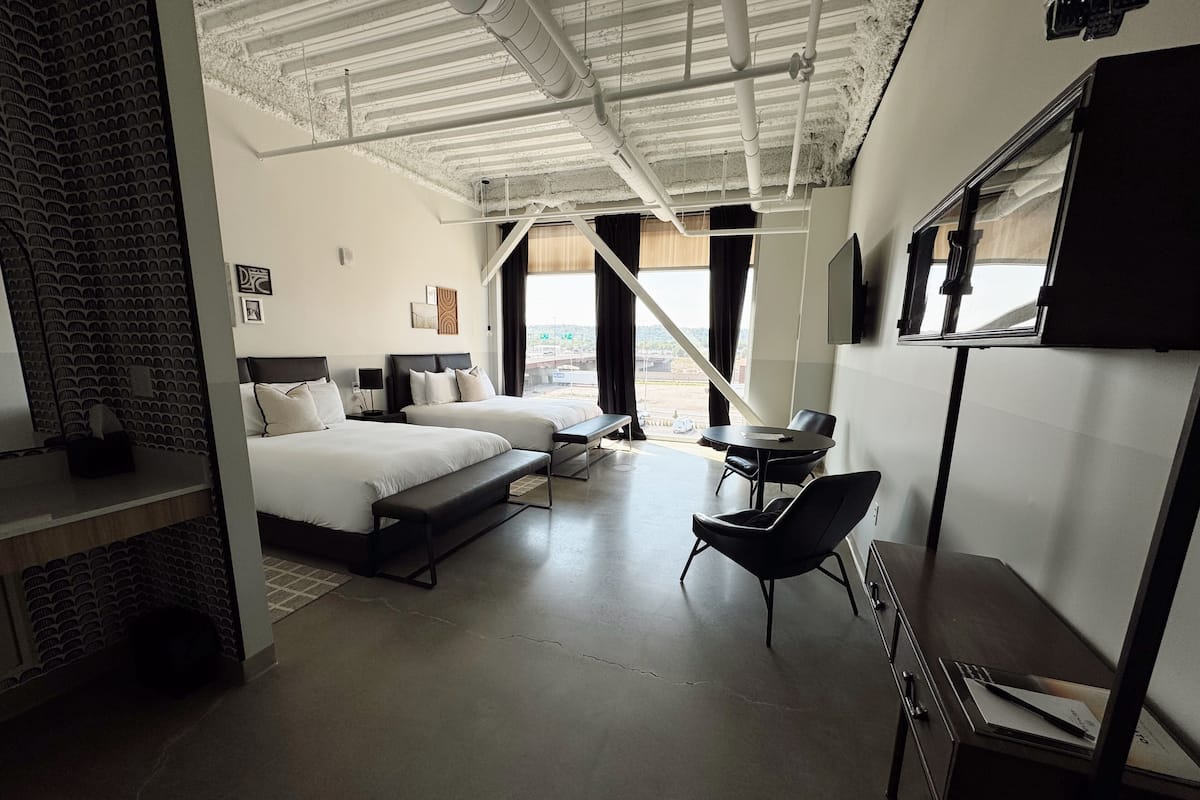A modern hotel room with two beds, a desk, and a large window offering a view of the city.
