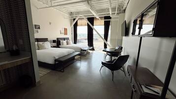 A modern hotel room with two beds, a desk, and a large window offering a view of the city.