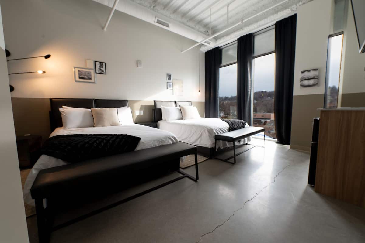 A modern hotel room with two beds, a bench, a desk, and large windows offering a city view.