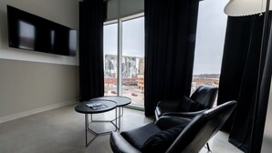 Een moderne hotelkamer met een groot raam, een flatscreen televisie aan de muur, een ronde tafel met een tijdschrift en een zwarte leren fauteuil.