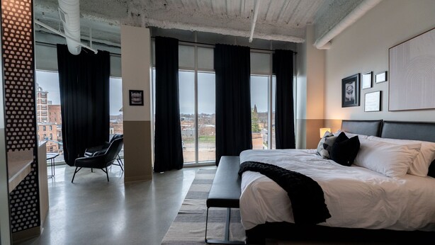 Een moderne hotelkamer met een groot bed, een bankje en uitzicht op de stad door grote ramen.
