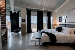 A modern hotel room with a large bed, a bench, and a view of the city through large windows. - Arch and Cable Hotel (Mankato)