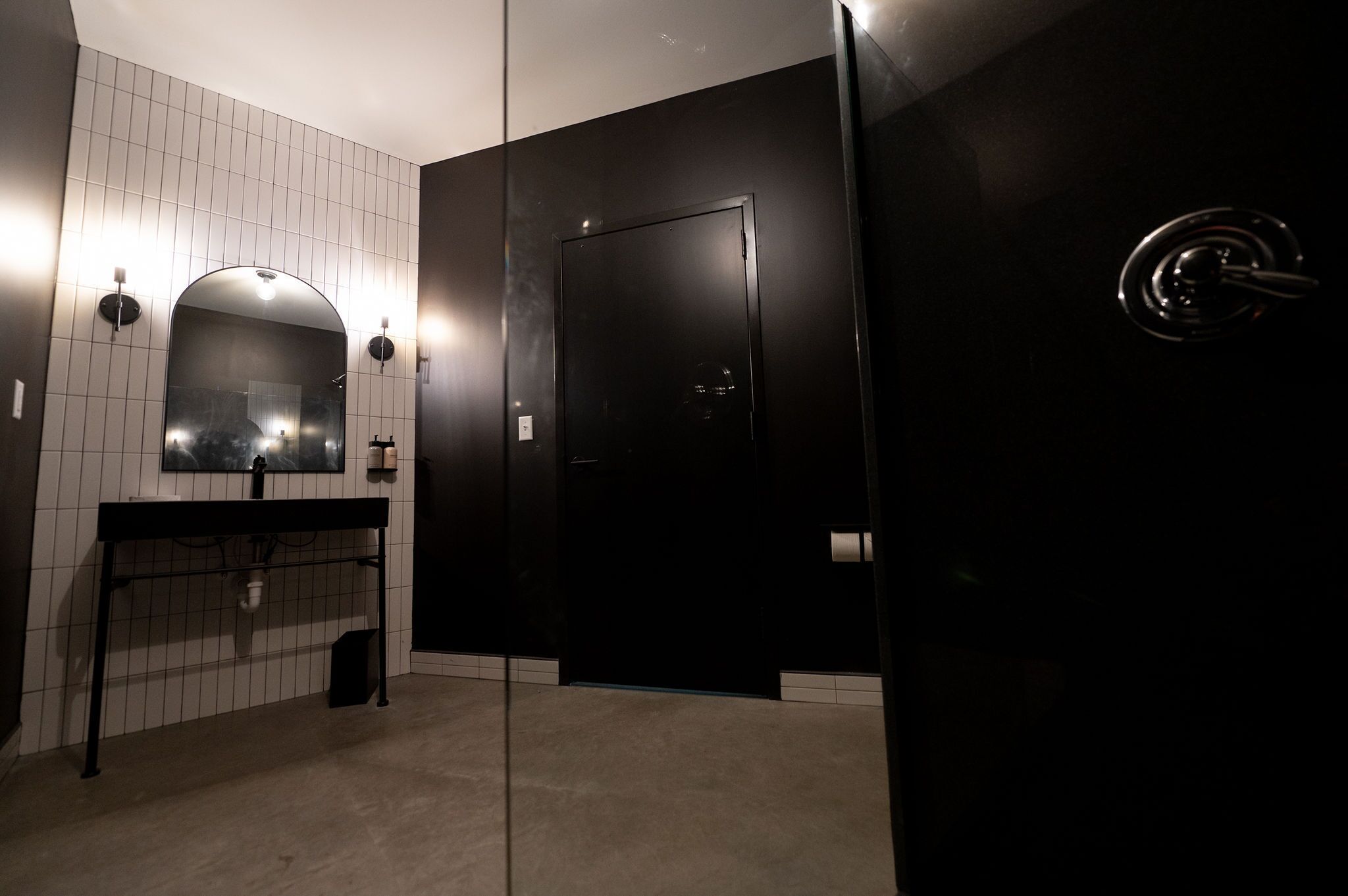 A modern bathroom with a black door, a white tiled wall, and a sink with a mirror.