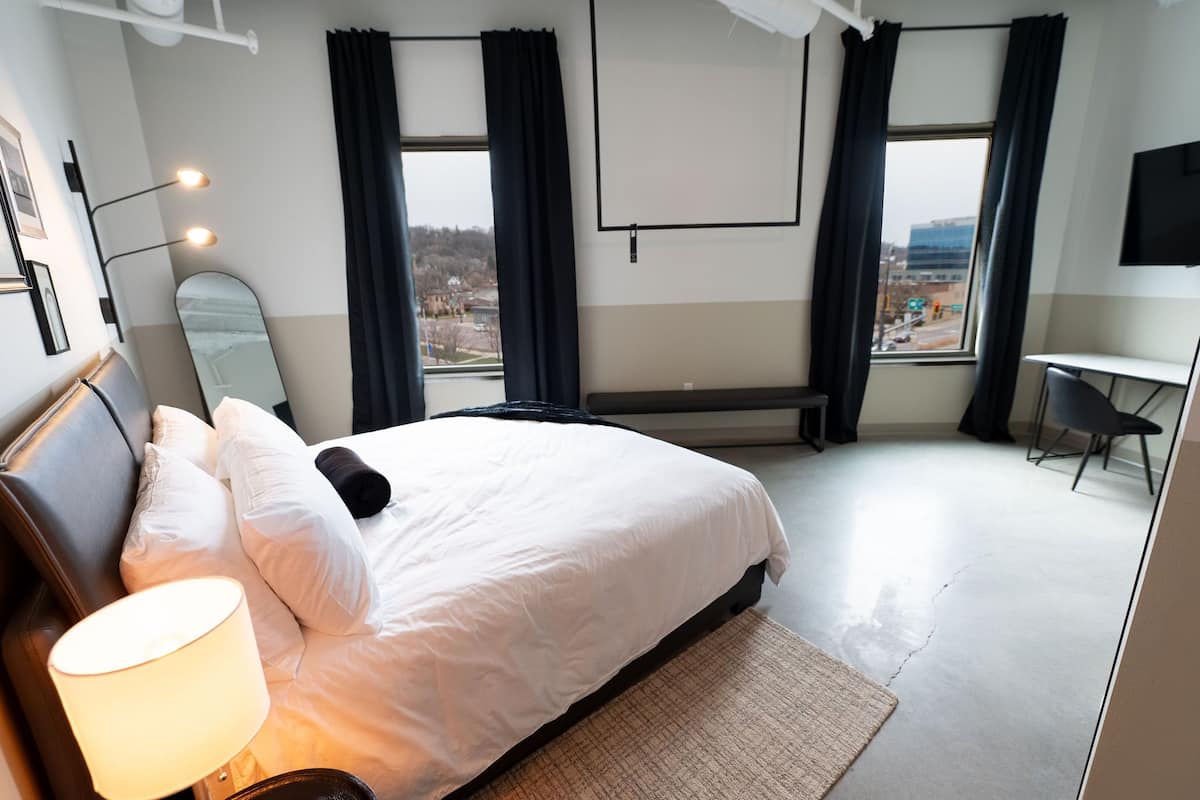A hotel room with a bed, a desk, a chair, a lamp, a mirror, and a view of the city through the window.
