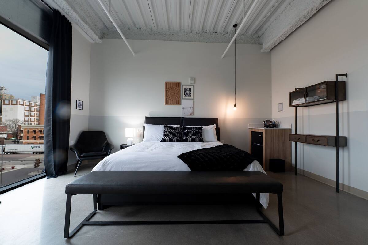 A modern hotel room with a large bed, a bench, a desk, and a view of the city.