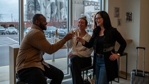 Drie personen proosten met champagnefluiten in een ruimte met grote ramen met uitzicht op een straat.