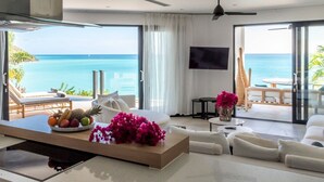 A modern living room with a large window overlooking the ocean, a flat-screen TV mounted on the wall, and a dining area with a fruit bowl and flowers. - Tamarind Hills Resort - Ocean Azul (Antigua and Barbuda)