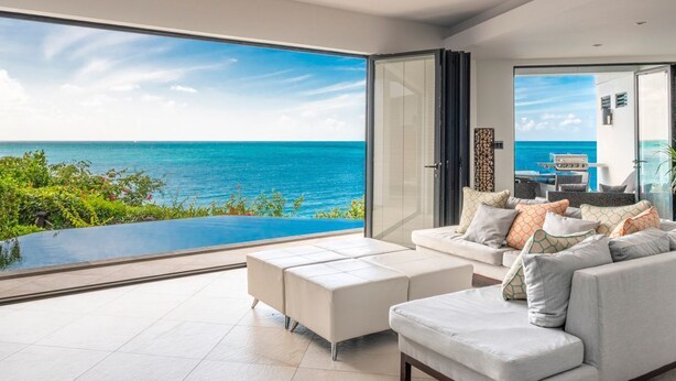 A modern living room with a large glass door leading to a balcony overlooking a pool and the ocean. - Tamarind Hills Resort - Ocean Blessed (Antigua and Barbuda)