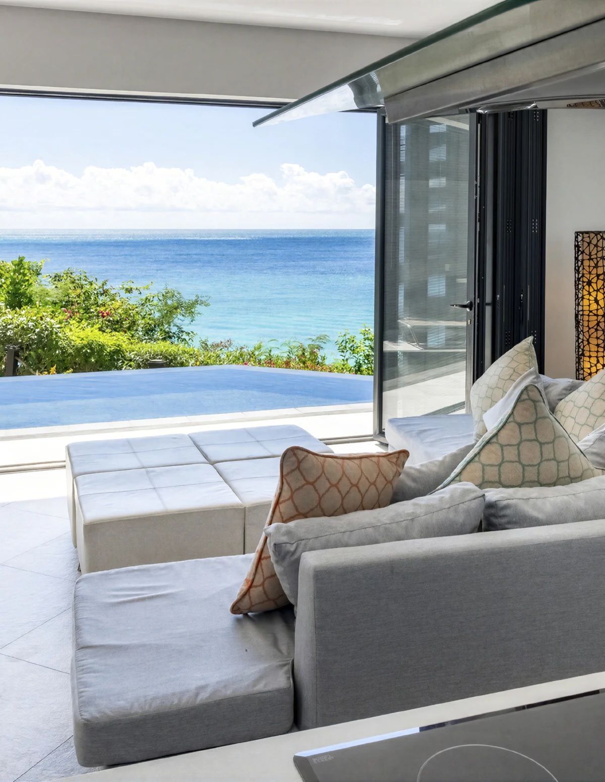 Un salon moderne avec un canapé et des coussins, une porte vitrée donnant sur un balcon avec vue sur l’océan.