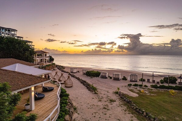 A beachfront property with a wooden deck, lounge chairs, and a view of the ocean at sunset.