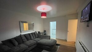A modern living room with a grey sectional sofa, a large mirror, a red pendant light, and a flat-screen TV mounted on the wall. - New! Xtra Large 4 Bedroom Up to 14 Guests (London)