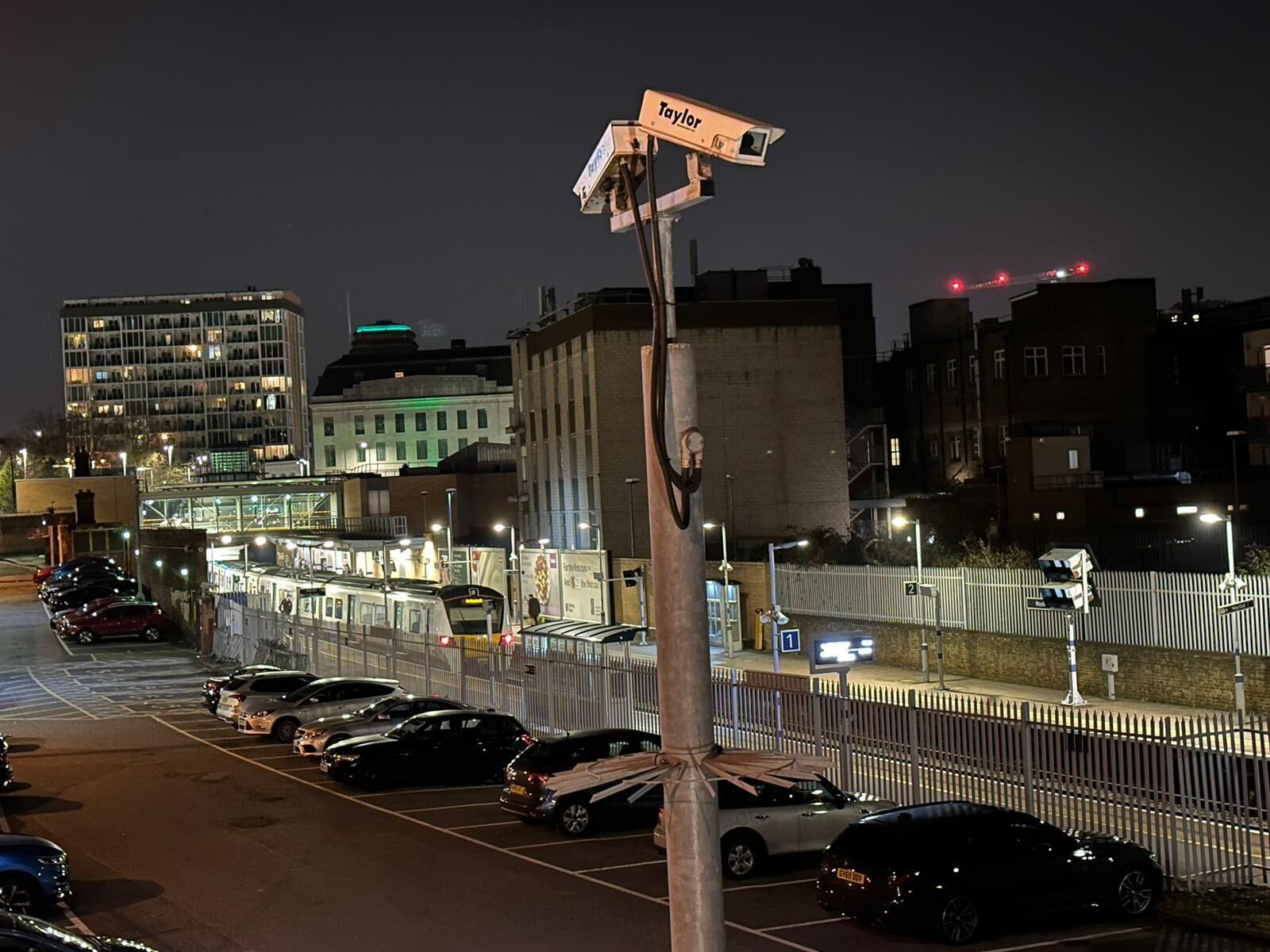 A nighttime cityscape with a parking lot, a tram, and buildings in the background.