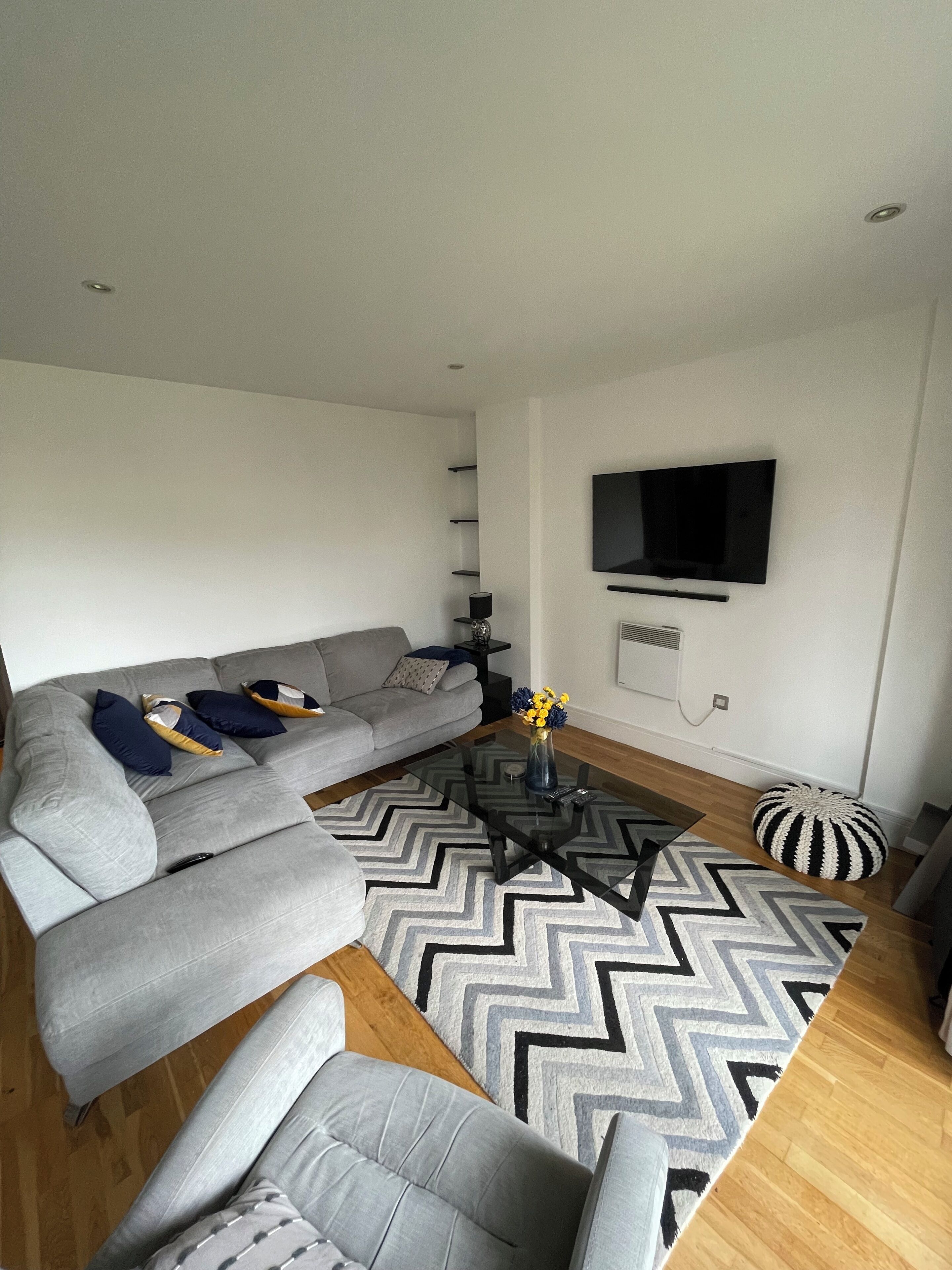 Un salon moderne avec un canapé d’angle gris, une table basse en verre, un téléviseur noir fixé au mur et un tapis à motifs.