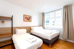 A hotel room with two beds, a wooden headboard, a window with curtains, and a painting on the wall. - Newly Renovated 1BD Flat, Heart of Camden (London)