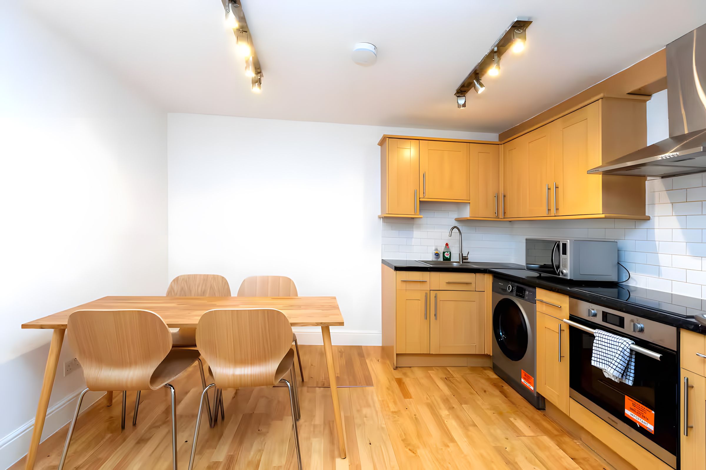 A modern kitchen with wooden cabinets, a dining table with chairs, a washing machine, and a microwave.