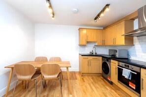 A modern kitchen with wooden cabinets, a dining table with chairs, a washing machine, and a microwave. - Newly Renovated 1BD Flat, Heart of Camden (London)