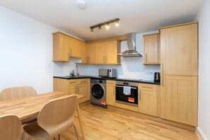 A modern kitchen with wooden cabinets, a washing machine, an oven, and a dining area with a table and chairs. - Newly Renovated 1BD Flat, Heart of Camden (London)