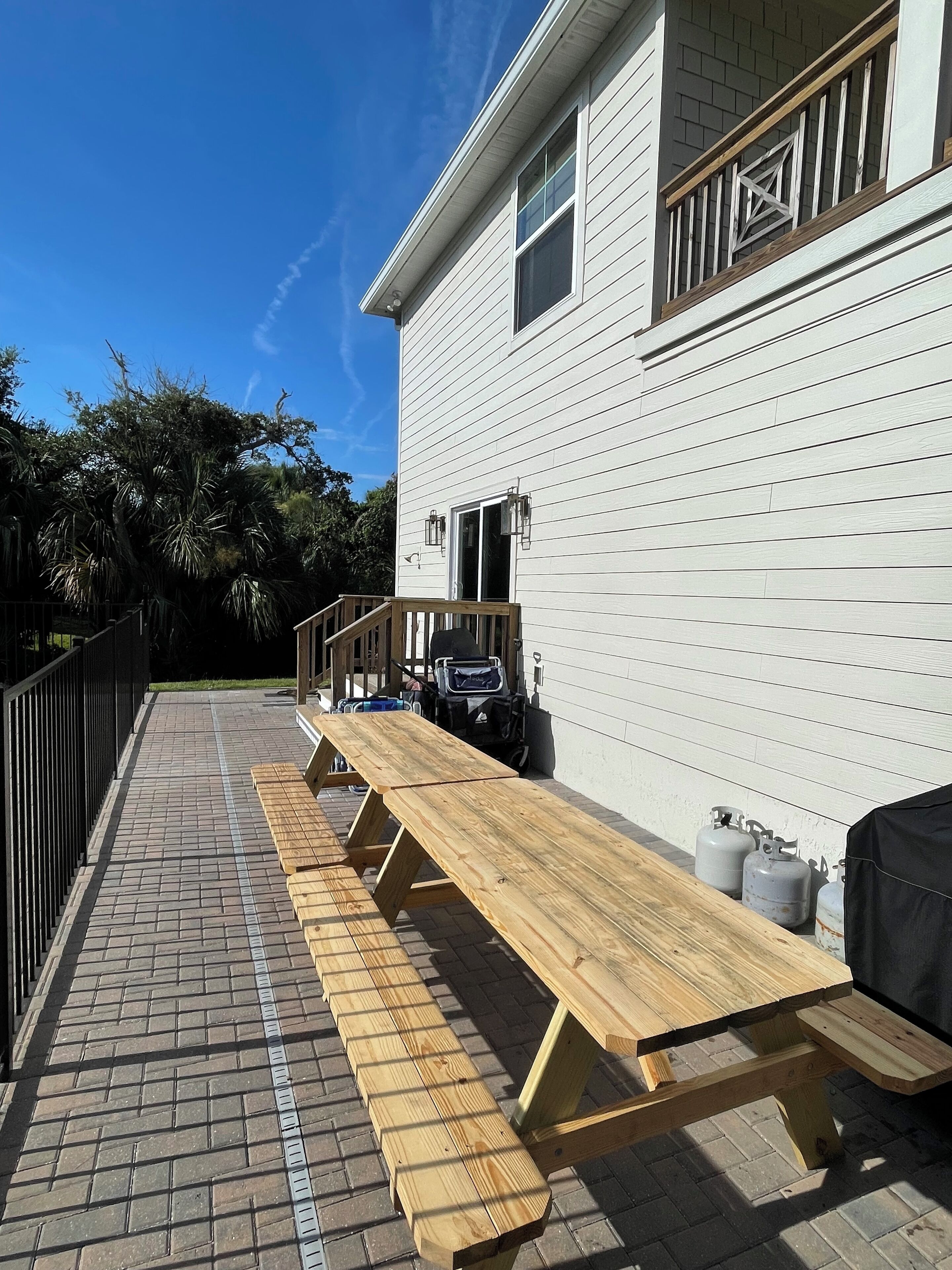 A wooden deck with picnic tables, a barbecue grill, and propane tanks against a white house with a balcony.