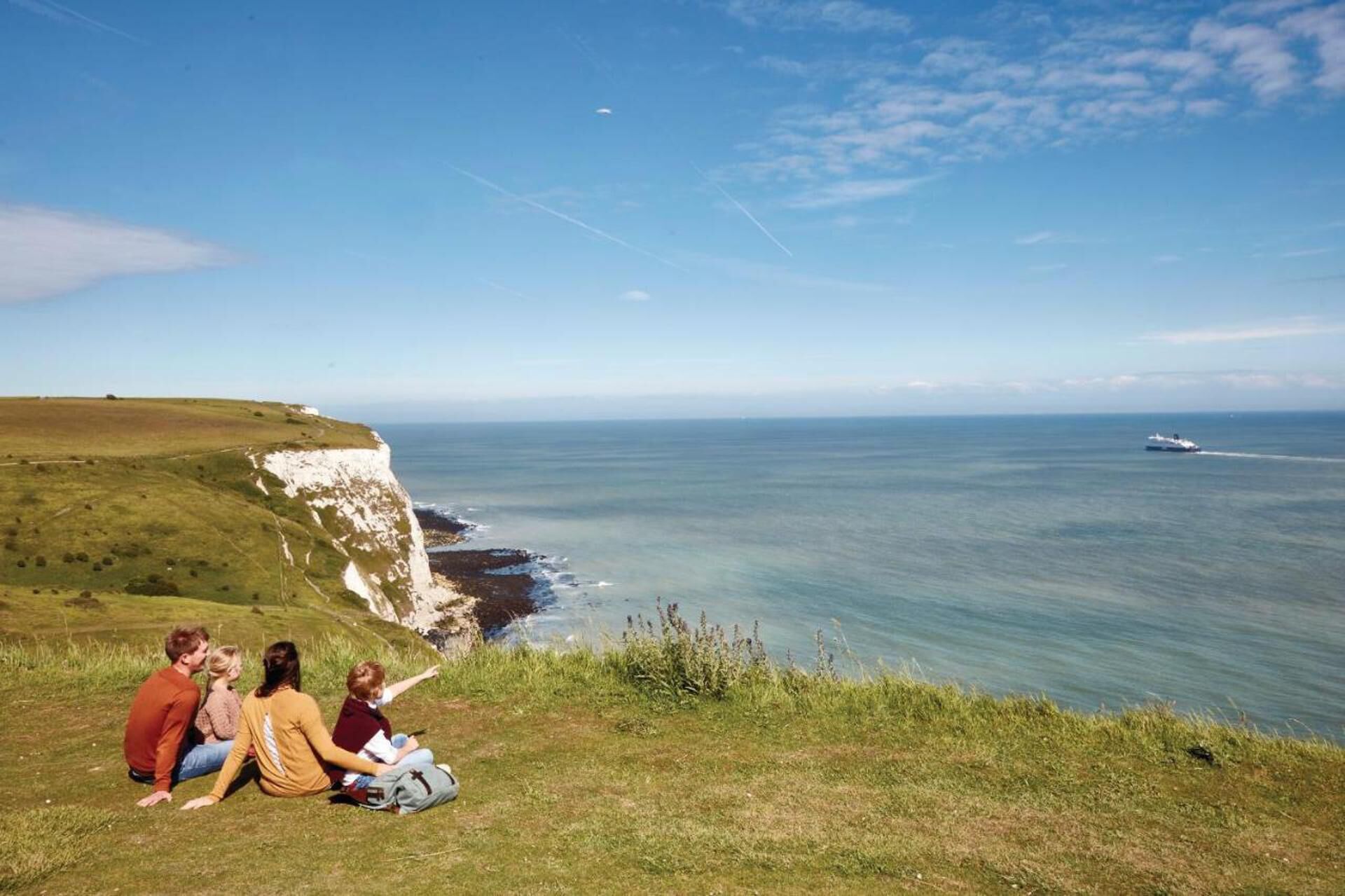 Un paysage côtier avec une falaise surplombant la mer, un bateau au loin et trois personnes assises dans l’herbe.