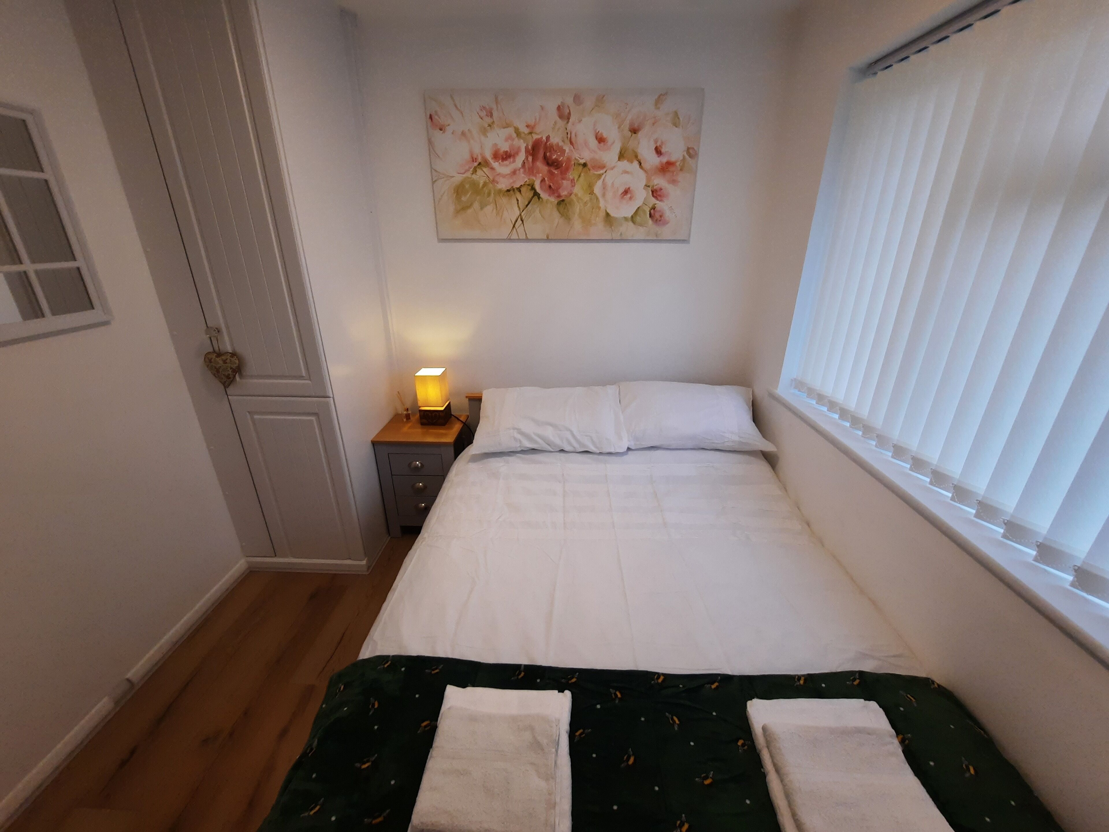 A neatly made bed with white linens, a green bedspread, and two towels. A small bedside table with a lamp is visible. A floral painting adorns the wall above the bed.