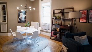 A dining area with a round table, white chairs, and a chandelier. There is a bookshelf with decorative items and a lamp.