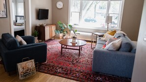 A living room with a blue sofa, a grey sofa, a coffee table, a red patterned rug, a television, and a window with a view of trees and a car outside.