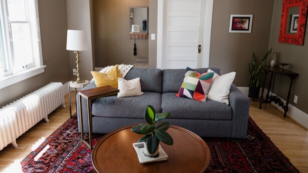 A living room with a grey sofa, a round wooden coffee table, a patterned rug, a radiator, and a window with blinds.