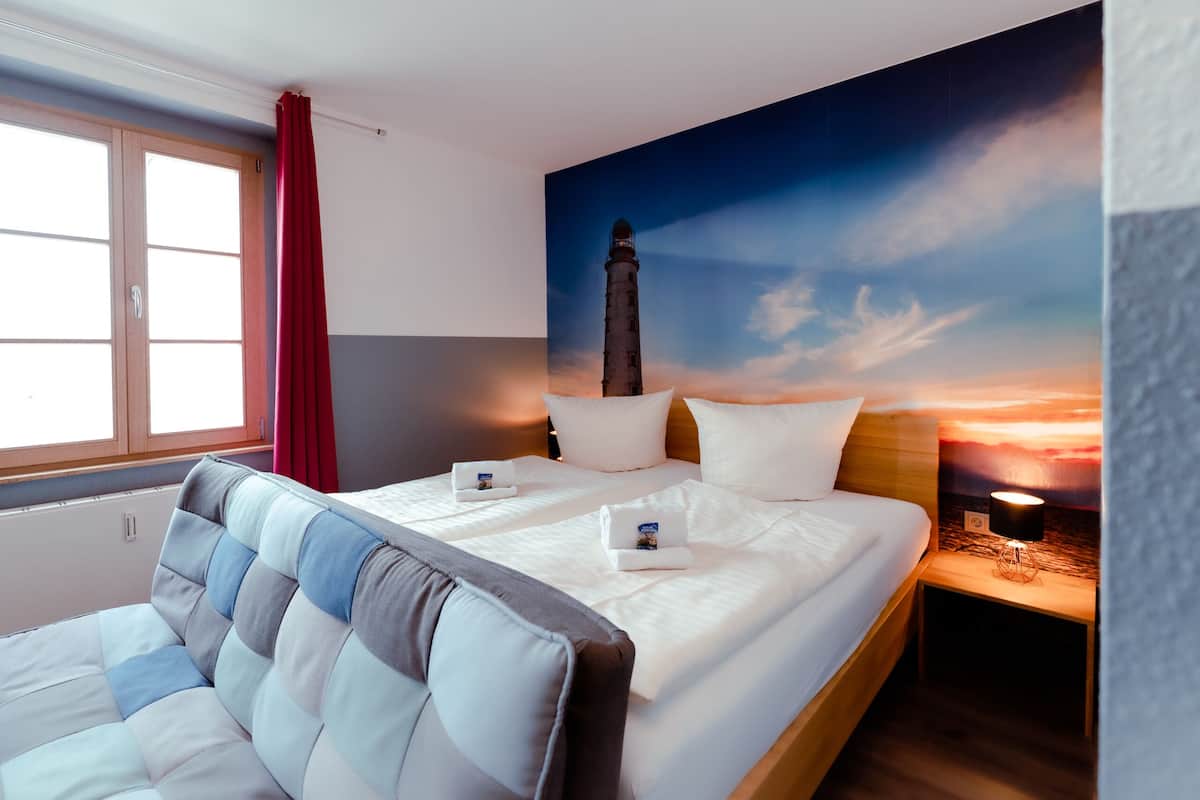 Et hotelværelse med en seng, et natbord, en lampe, et vindue med gardiner og et vægmaleri af et fyrtårn.