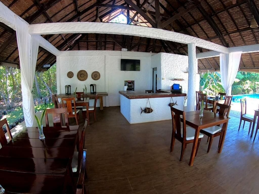 A covered outdoor dining area with wooden tables and chairs, a central bar area, and a view of the surrounding greenery.
