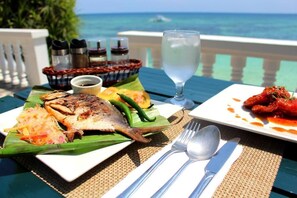 A table set with a grilled fish, a plate of red peppers, and a glass of water, with a view of the ocean in the background. - Casa Amihan (Anda)