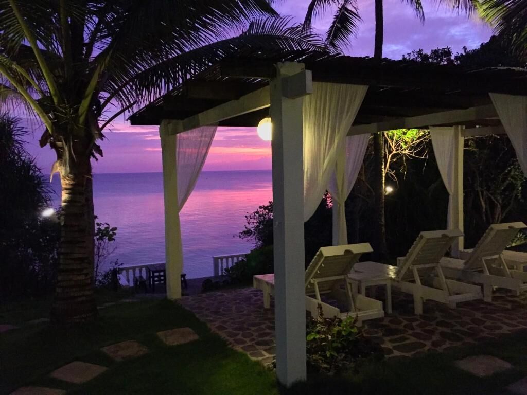 A covered outdoor seating area with lounge chairs, a view of the ocean, and a sunset in the background.