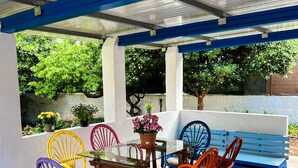 Une terrasse couverte avec une table en verre et des fauteuils en osier aux couleurs vives, le tout entouré de verdure.