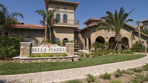 A multi-story building with a stone facade, a sign reading "Bella Piazza," and a landscaped entrance with palm trees.