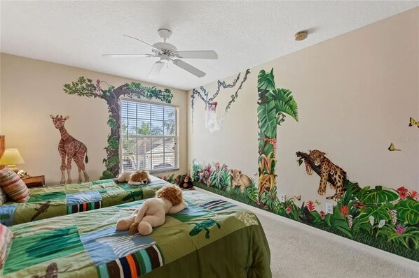 Une chambre avec deux lits, un ventilateur de plafond et une fresque représentant des animaux de la jungle sur le mur.