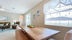 Un salon spacieux avec une table à manger en bois, deux canapés et une table basse. Il y a deux fenêtres équipées de stores, un ventilateur de plafond et des œuvres d’art accrochées aux murs.