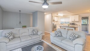 Un salon moderne avec deux canapés gris, un ventilateur de plafond et une cuisine en arrière-plan.