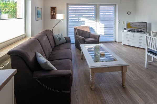 A living room with a brown leather sofa, a glass-top coffee table, a white side table, a white dresser, and a television on the dresser. - Ferienhaus Norden - Berliner Str. 24 offers you plenty of space and comfort for a relaxing vacation in a quiet location. Look forward to cozy accommodation with good connections to the city center, excursion destinations and the North Sea coast. (Norden)
