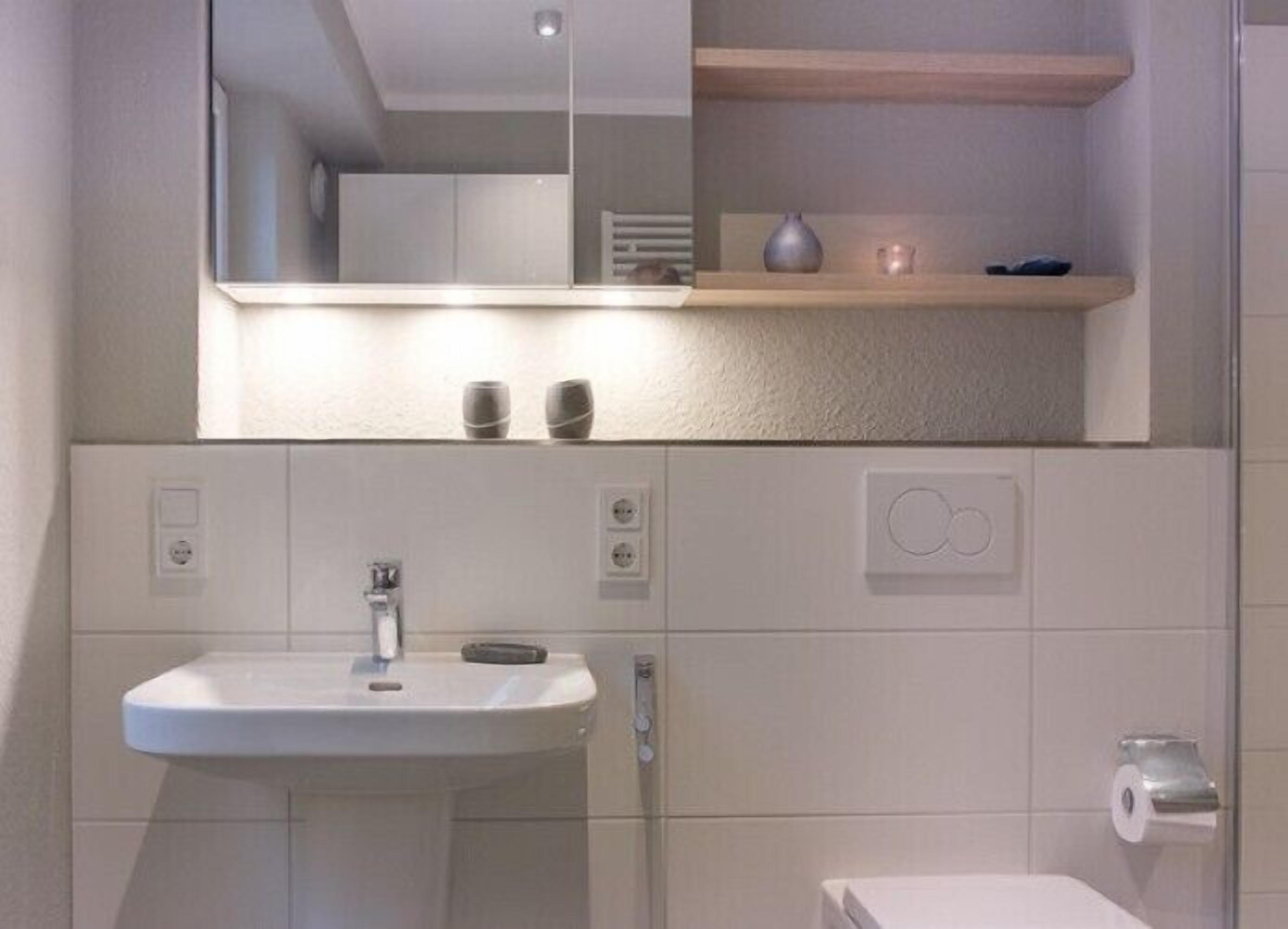Un baño moderno con un lavamanos, espejo y retrete blancos. Hay dos repisas sobre el lavamanos con objetos decorativos.