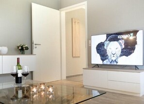 Un salon moderne doté d’un téléviseur à écran plat affichant une image, d’une table basse en verre ornée de bougies et d’une bouteille de vin.
