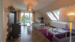 A modern living room with a wood-burning stove, a dining area, a TV, and a large window with a view of the outdoors.