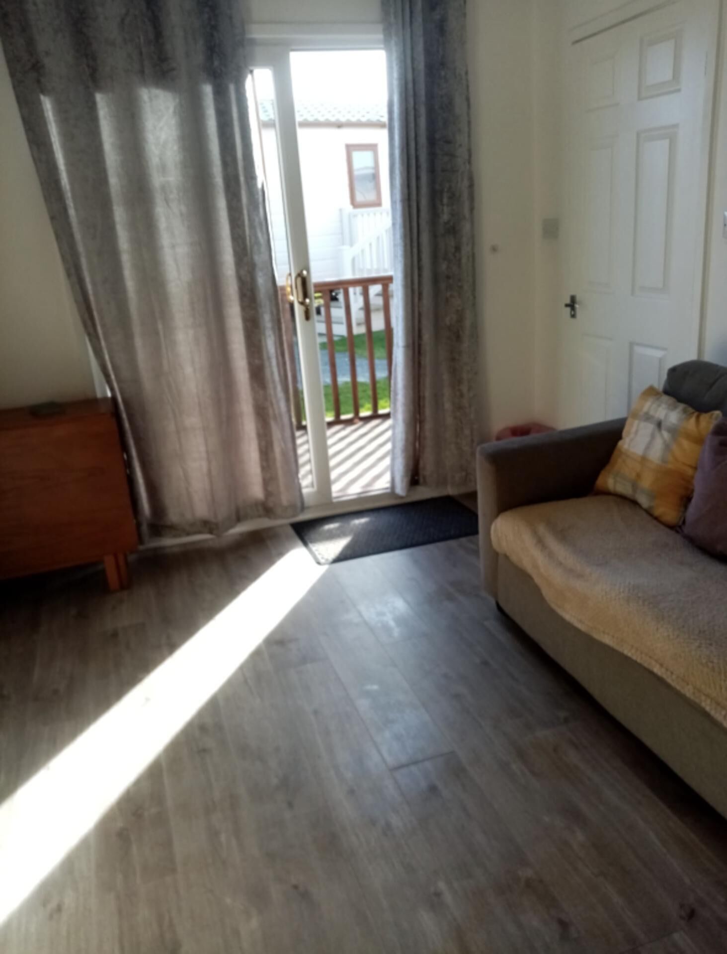 A room with a wooden floor, a sofa with a blanket, a wooden side table, and a sliding glass door leading to an outdoor area with a railing.