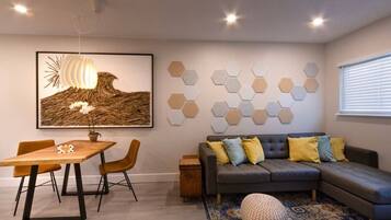 Un salon moderne comprenant un canapé d’angle, une table à manger avec des chaises, une table basse, un grand tableau abstrait, des éléments de décoration murale hexagonaux et un tapis à motifs.