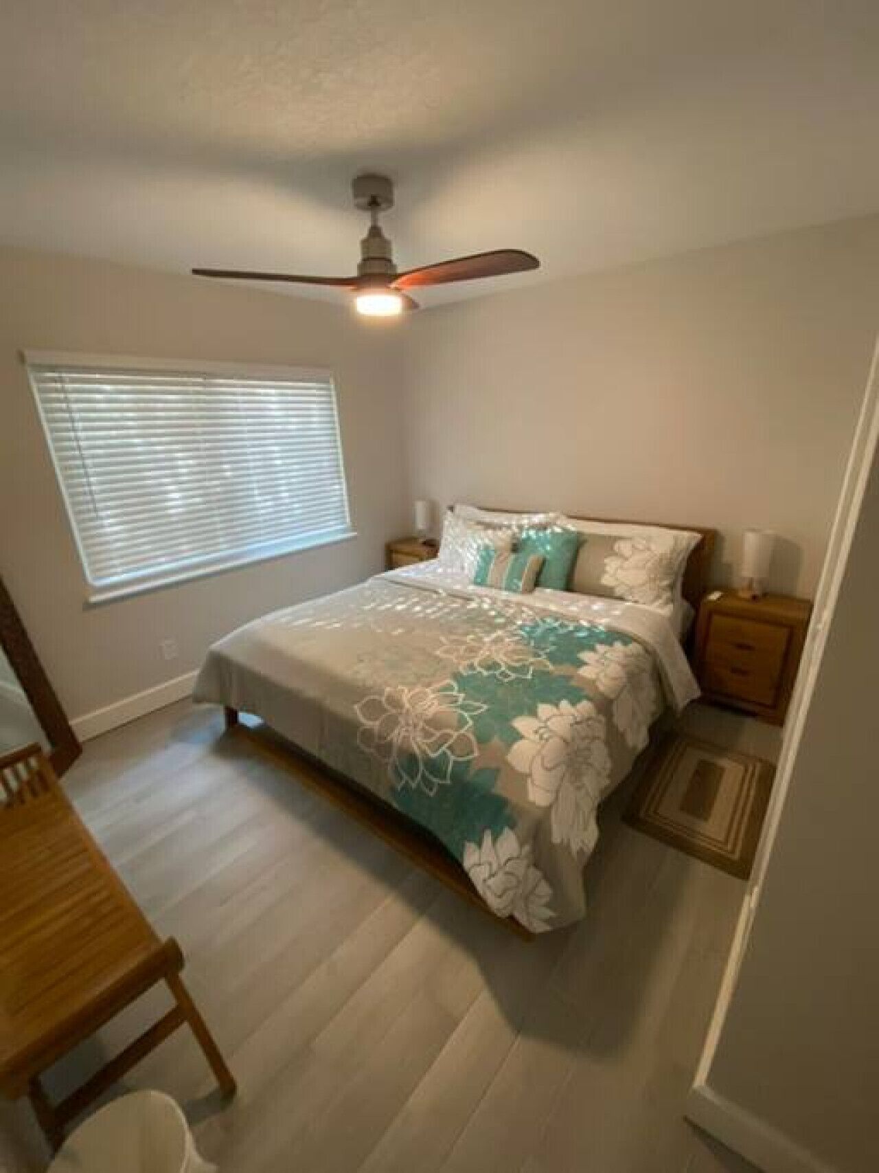 Une chambre à coucher comprenant un lit, un ventilateur de plafond, une fenêtre avec des stores, une table de chevet et une commode.