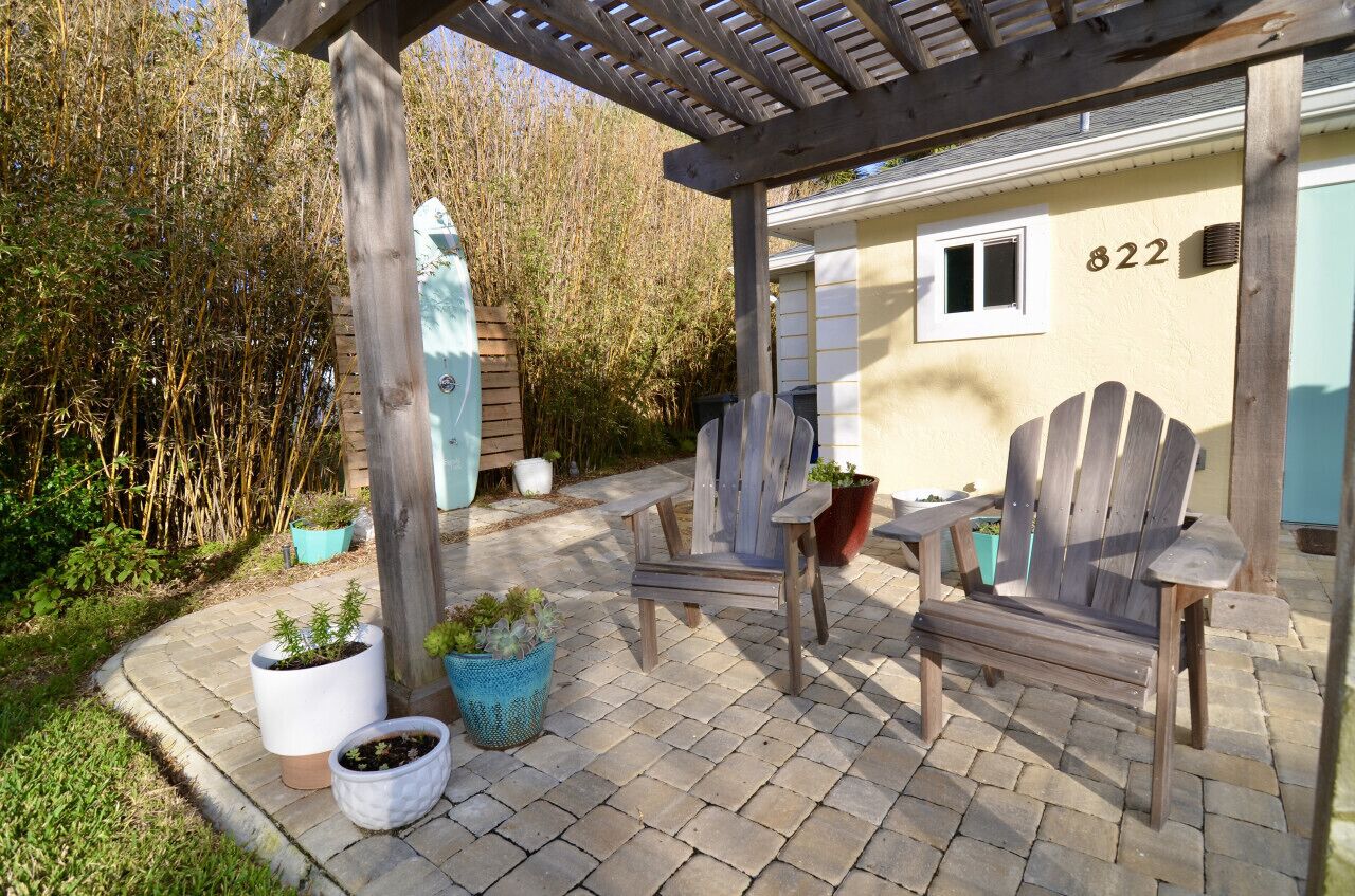 Een houten pergola met een bankje en potplanten, een huis met nummer 822, en een bamboomschutting op de achtergrond.