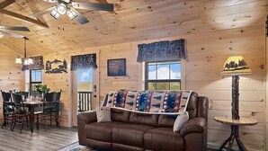 Ein gemütliches Blockhauskamin-Interieur mit einer braunen Ledersofa, Holzwänden, einem Essbereich mit Stühlen und einem Deckenventilator.