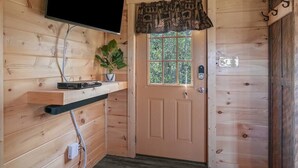 Ein Holzblockhaus-Interieur mit einem angebrachten Fernseher, einer Tür mit gemustertem Vorhang, einem Blumentopf und einem Regal mit elektronischen Geräten.