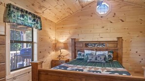 Ein Schlafzimmer einer Holzhütte mit Bett, Nachttisch, Lampe und Fenster mit Blick ins Freie.