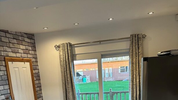 A room with a sofa, a wooden table, a refrigerator, and a sliding glass door leading to a balcony with a view of a green lawn and a building. - Lovely 1-bed Chalet in Mablethorpe (Mablethorpe)