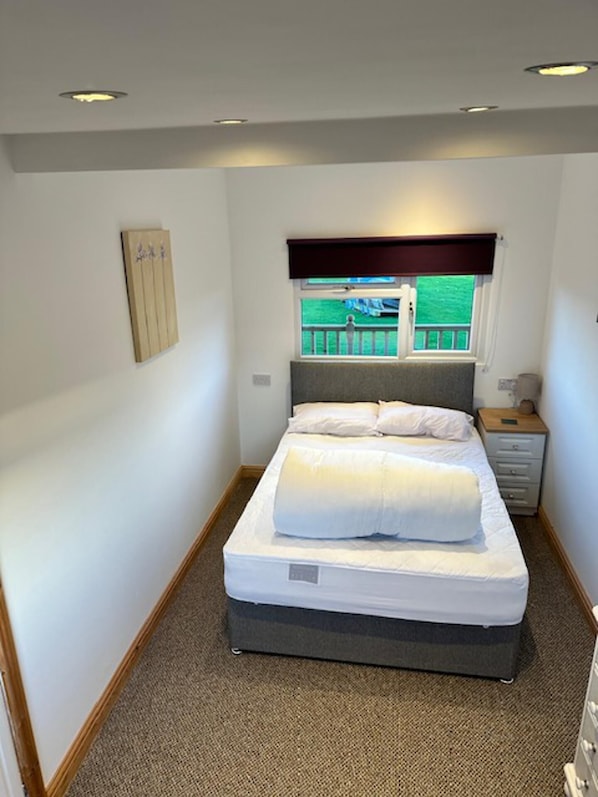 A single bed with a headboard, a bedside table, a nightstand, and a window with a view of greenery outside.